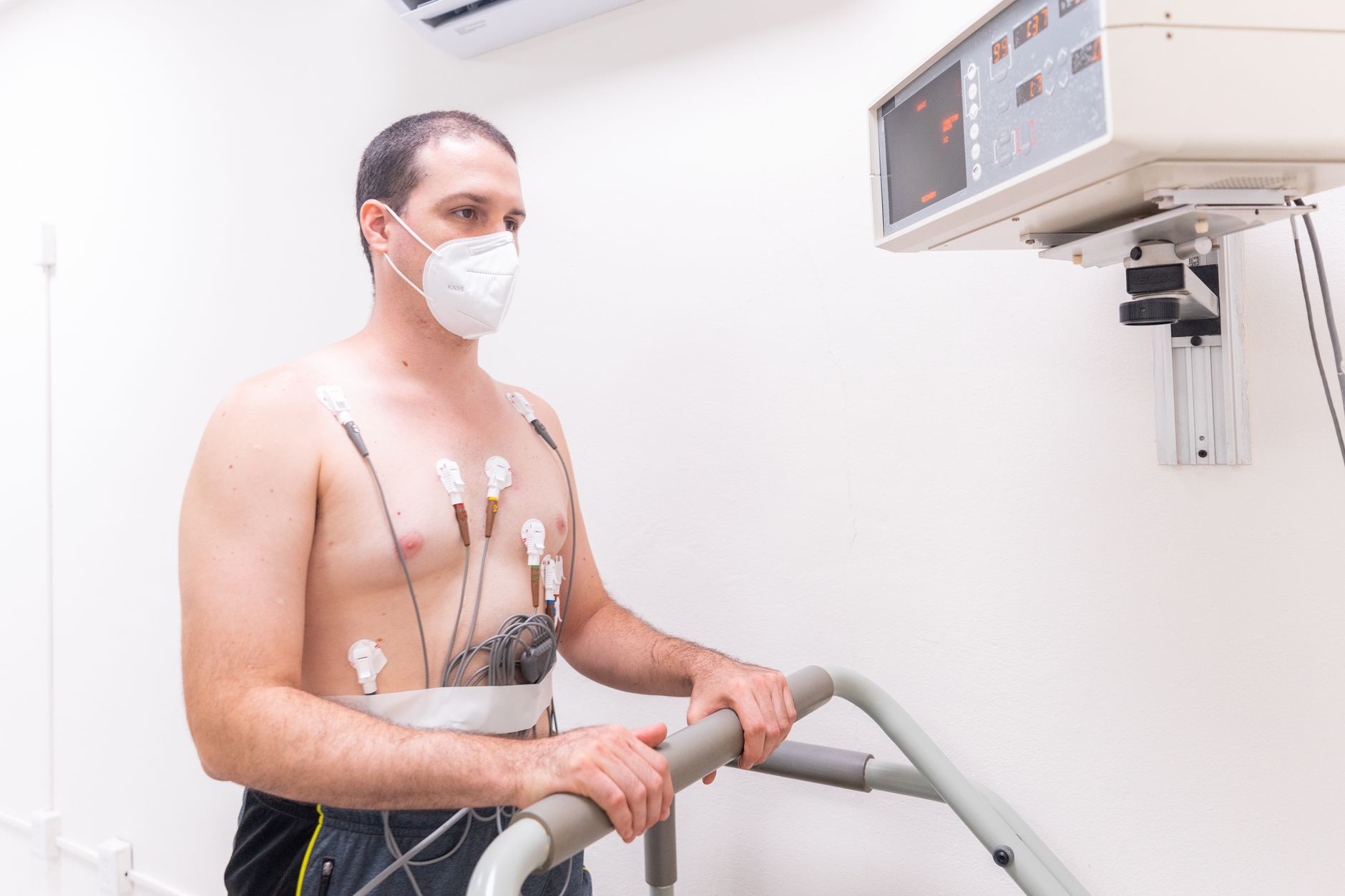 A patient on a treadmill wearing chest electrodes and a gas-exchange mask during a graded exercise test, with clinicians watching the readout