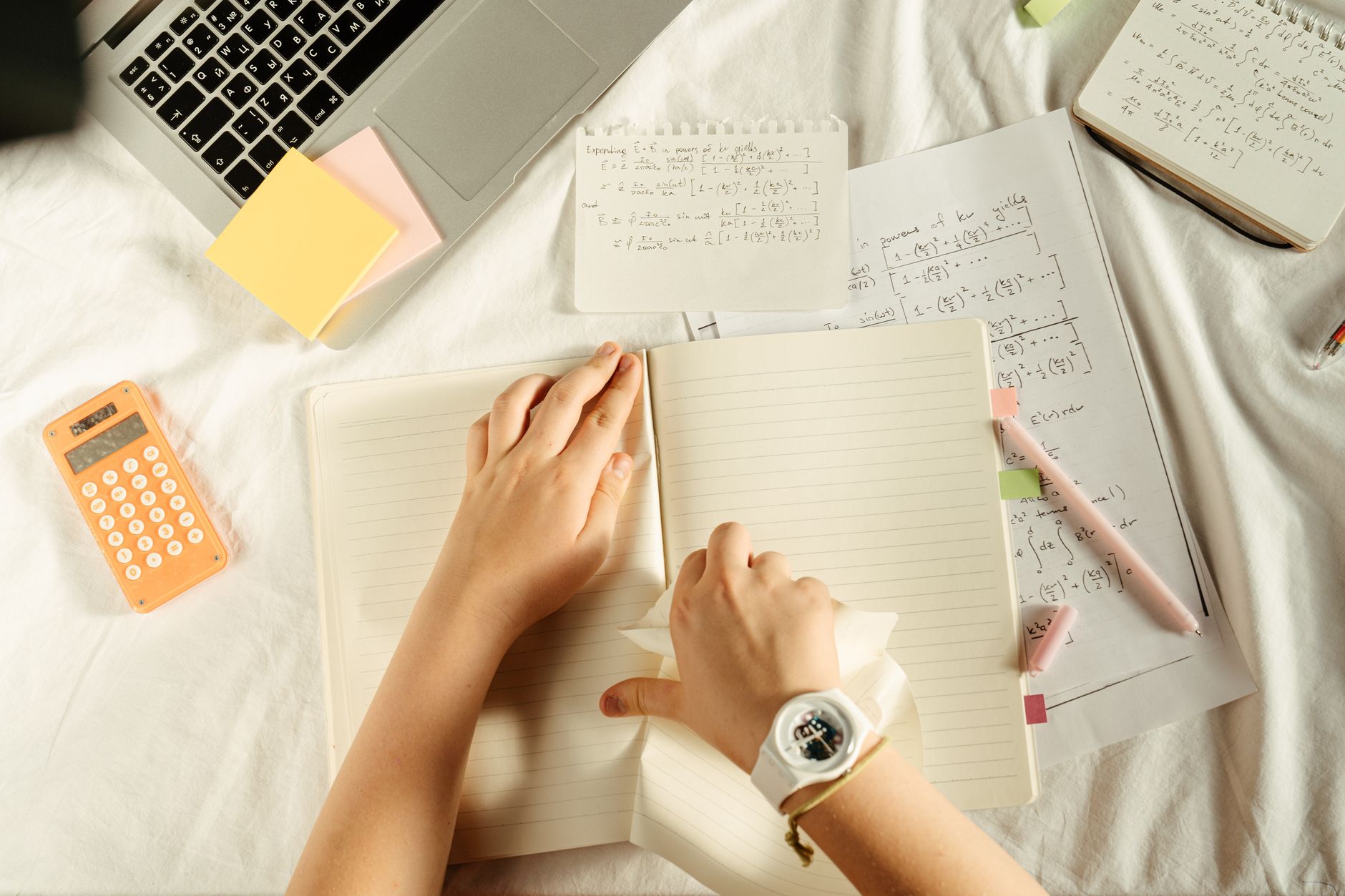 Overhead view of a desk with a laptop, an orange calculator and open notebooks while a student tears out a page