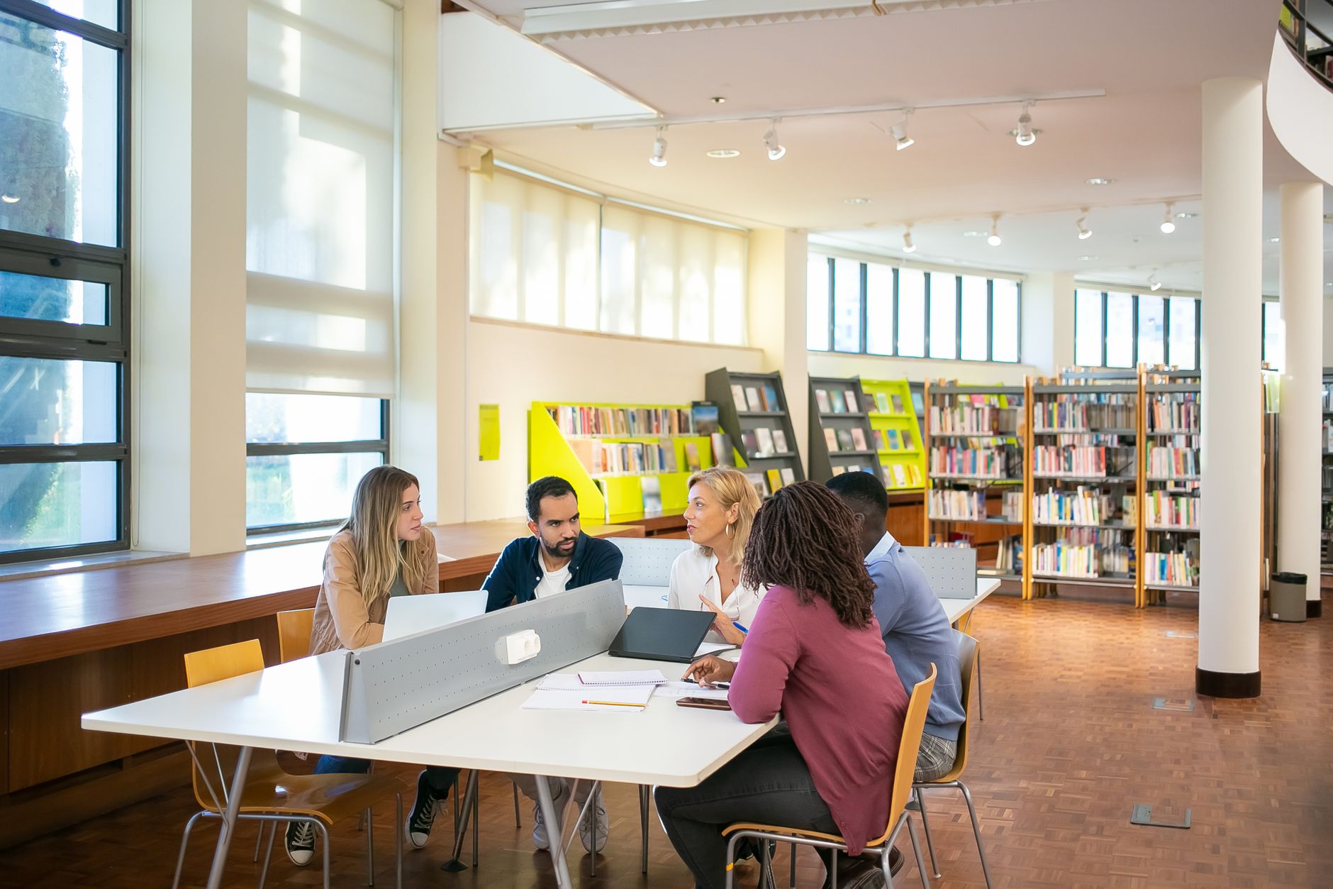 Five graduate students around a shared library table with laptops and notebooks, mid-discussion