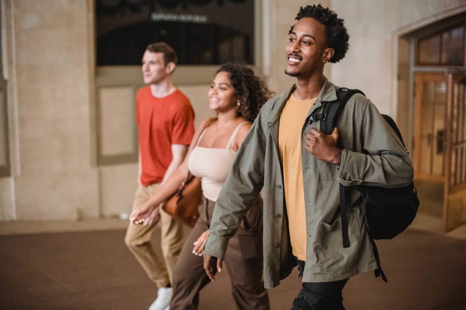 Three smiling university students walking together through a stone campus corridor, one carrying a backpack