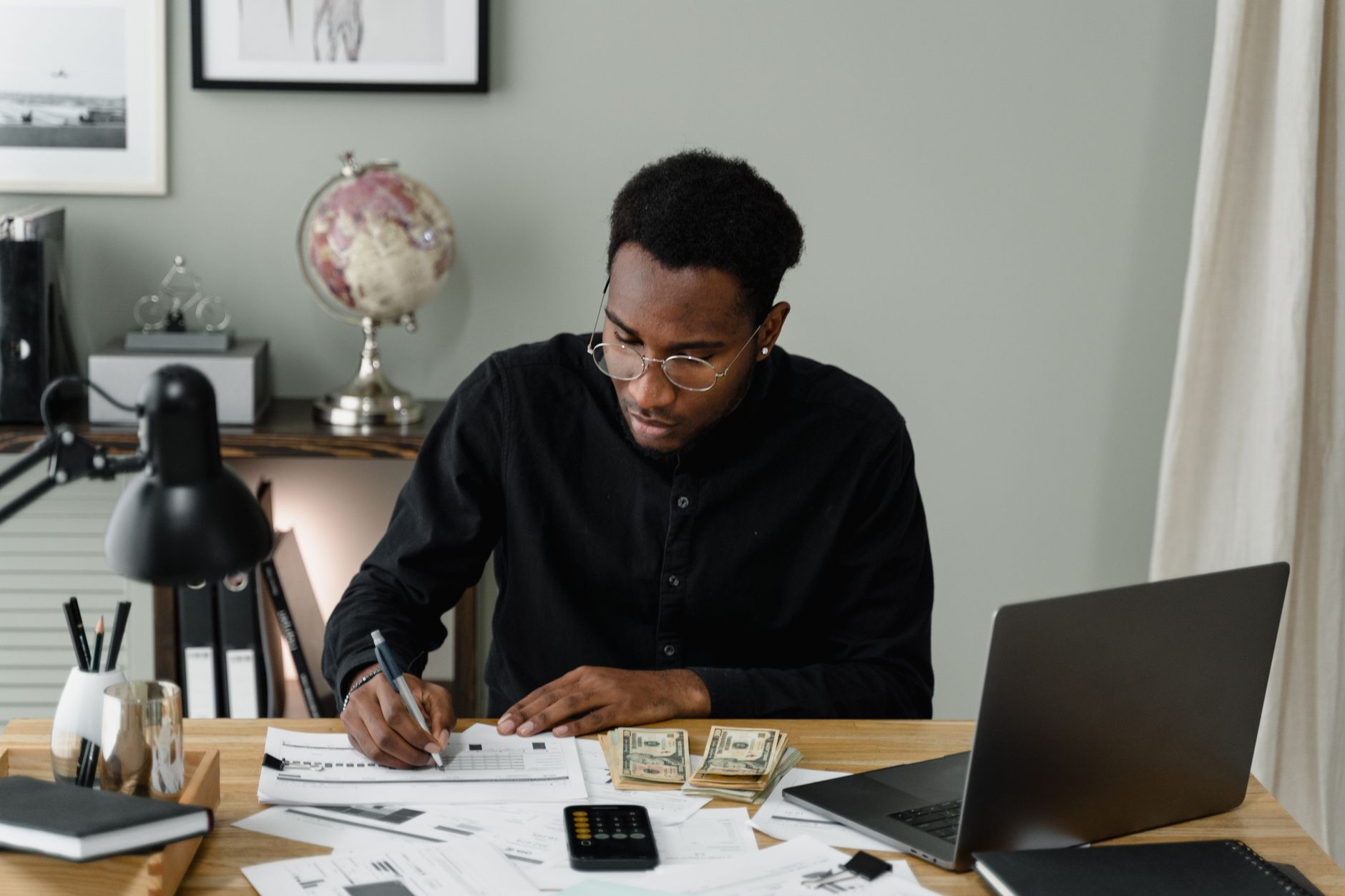 A man in glasses at a home desk writing in a budget sheet beside cash, a calculator and an open laptop