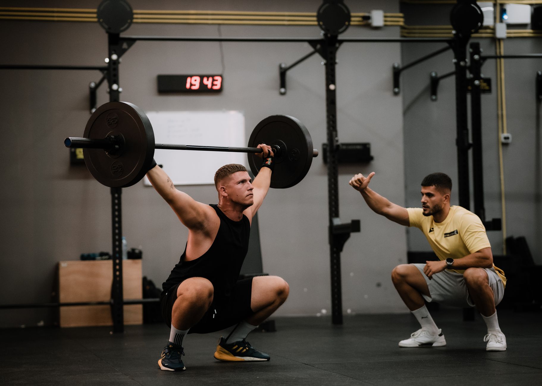 A coach guiding an athlete through a loaded barbell squat in a weight room